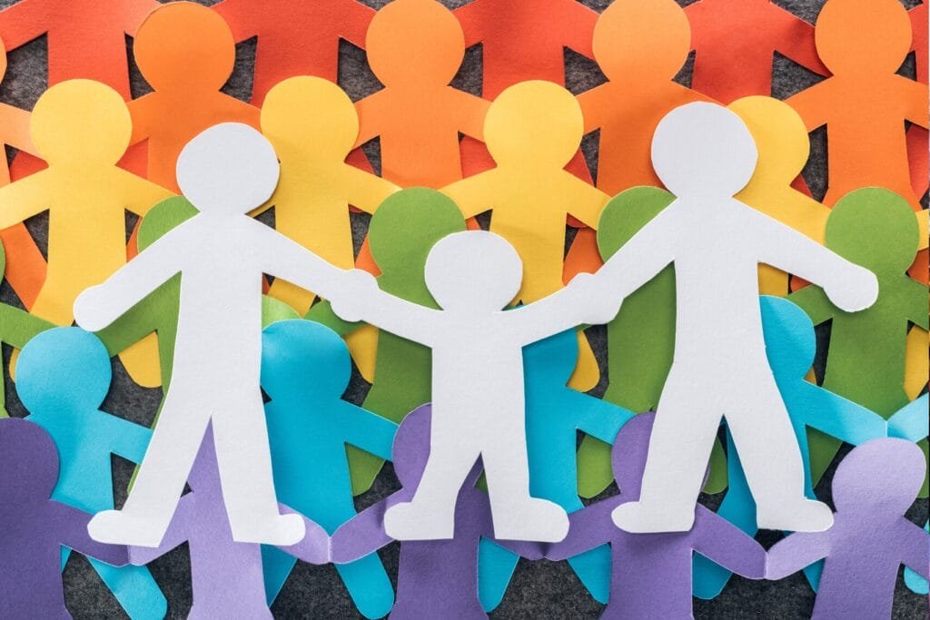 Colorful paper cutout figures forming a rainbow backdrop behind a white paper family holding hands, symbolizing LGBTQ+ family inclusion, unity, and diversity.