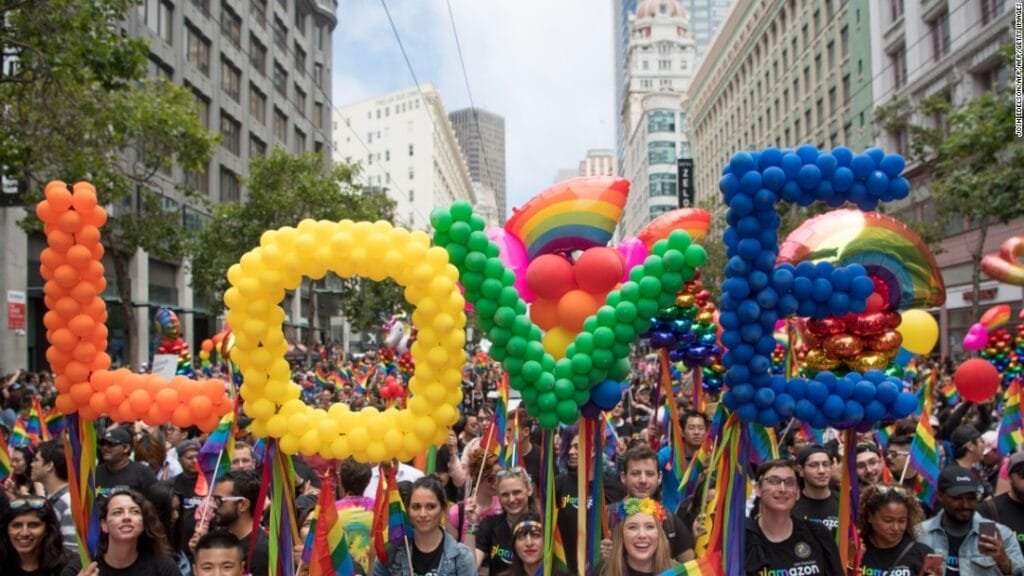 Participants in a vibrant Pride parade holding large rainbow balloon letters spelling “LOVE” amid a joyful crowd in downtown San Francisco.