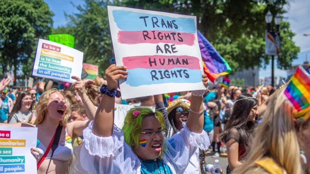 Transgender rights protestors marching at a Pride rally, holding up a hand-painted sign reading “Trans Rights Are Human Rights,” surrounded by rainbow flags and supportive messages.