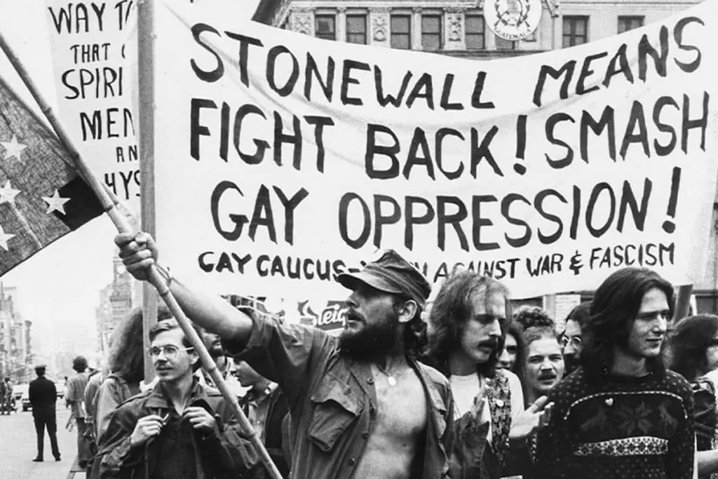 Black-and-white photo of protestors holding a “Stonewall means fight back! Smash gay oppression!” banner during an early LGBT rights demonstration.