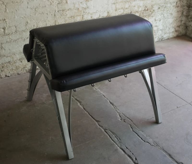 A padded black spanking bench from Stockroom.com, with metal legs designed for comfort, restraint, and supported positioning.