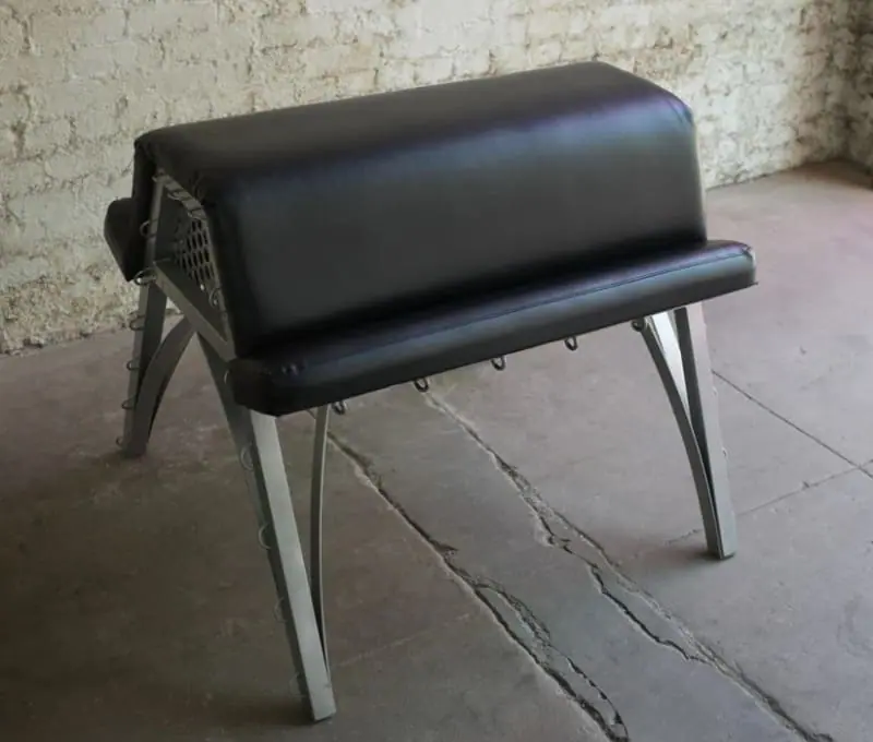A padded black spanking bench from Stockroom.com, with metal legs designed for comfort, restraint, and supported positioning.