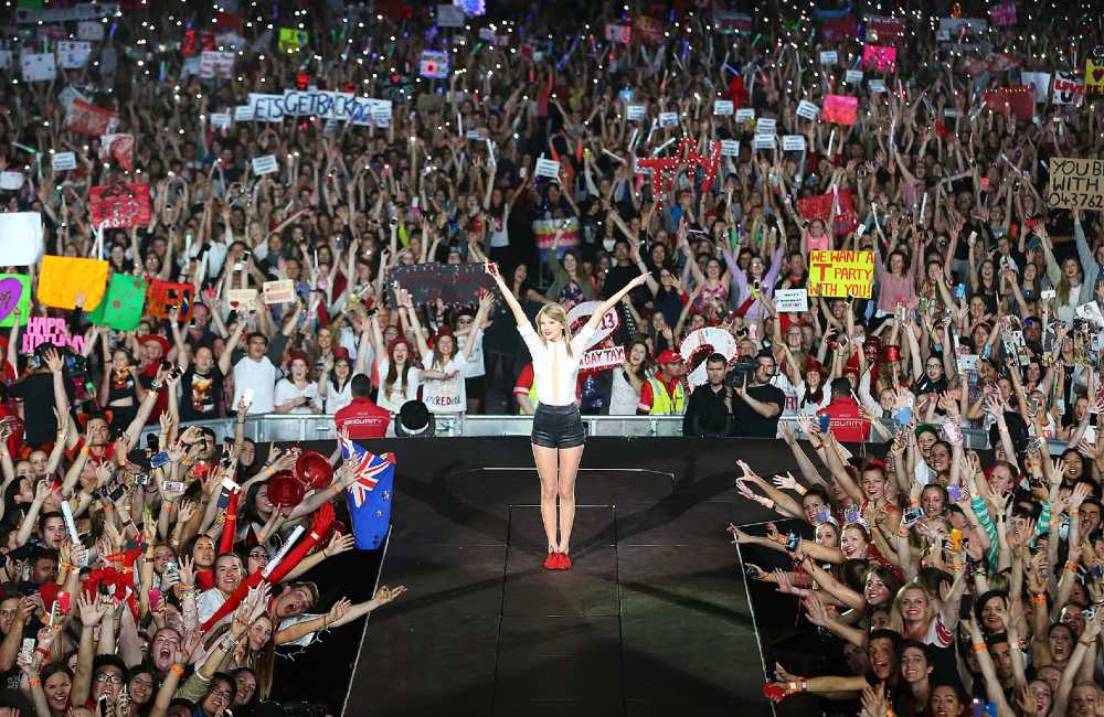 Taylor Swift stands on stage with her arms raised as thousands of fans cheer and hold up signs during the Eras Tour, symbolizing the power of the Swiftie community.
